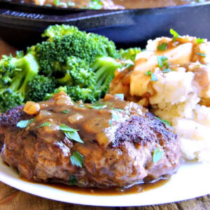 Closeup of a salisbury steak on a white plate topped with caramelized onion gravy and accompanied by steamed broccoli and mashed potatoes.