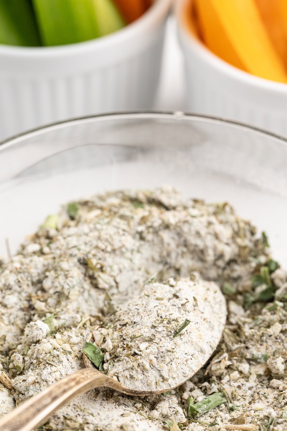 Close-up of the best homemade ranch dressing mix in a glass bowl on a white counter with white ramekins full of fresh vegetable sticks in the background.