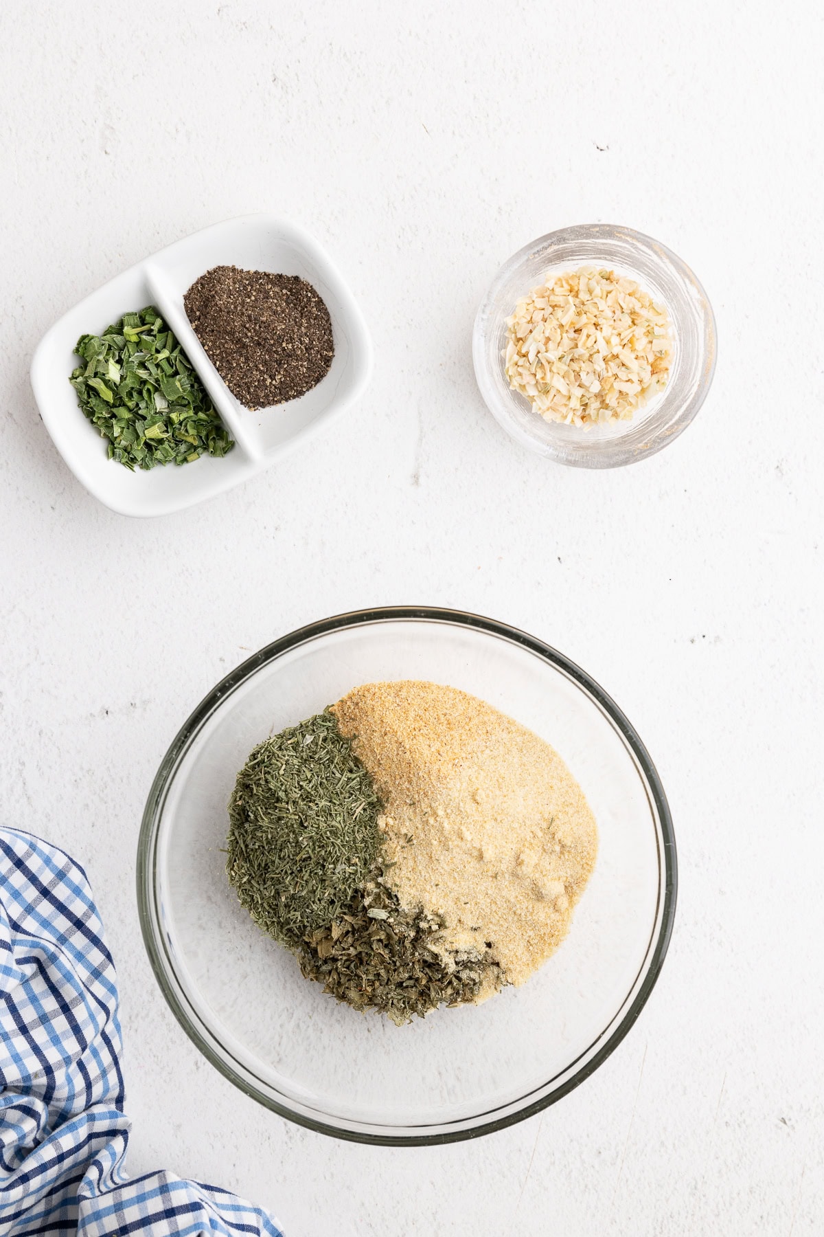 Overhead view of dried parsley, dill, garlic powder, and onion powder in a small glass bowl with minced onion, dried chives, and ground black pepper in small bowls next to it.