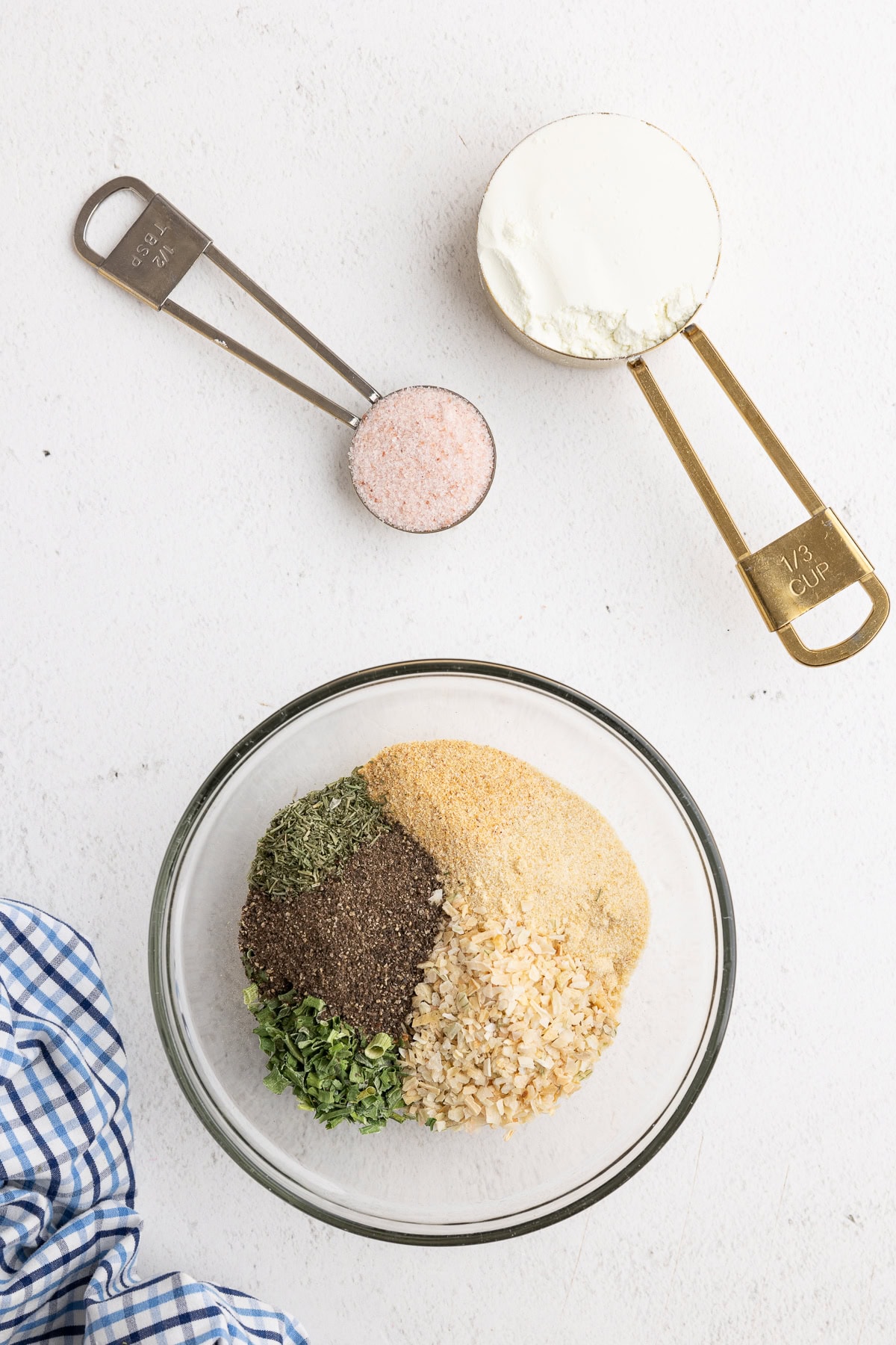 Overhead view of dried parsley, dill, chives, black pepper, onion powder, garlic powder, and dried minced onion in a small glass bowl with a measuring cup of buttermilk powder and a measuring spoon of pink salt next to it on a white counter.