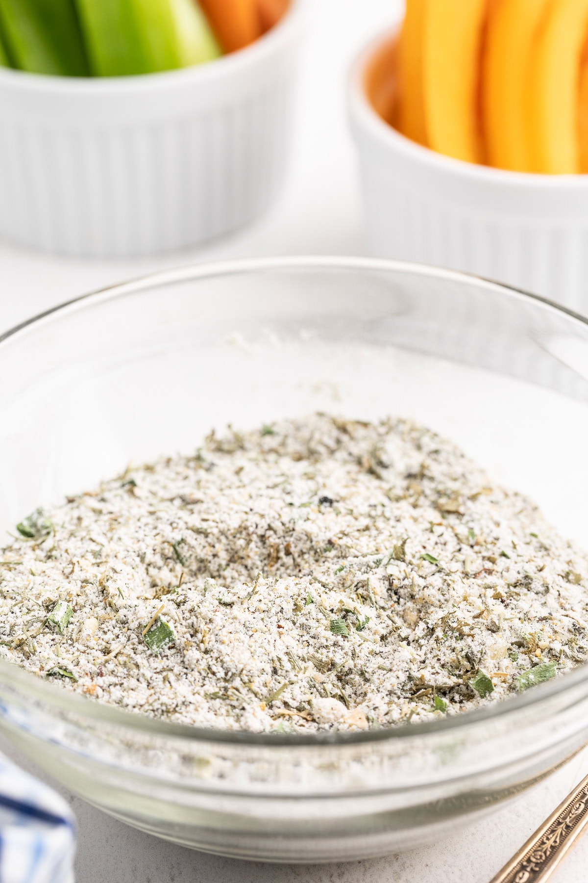 Overhead view of homemade ranch seasoning in a glass bowl on a white counter with ramekins full of fresh veggie sticks.