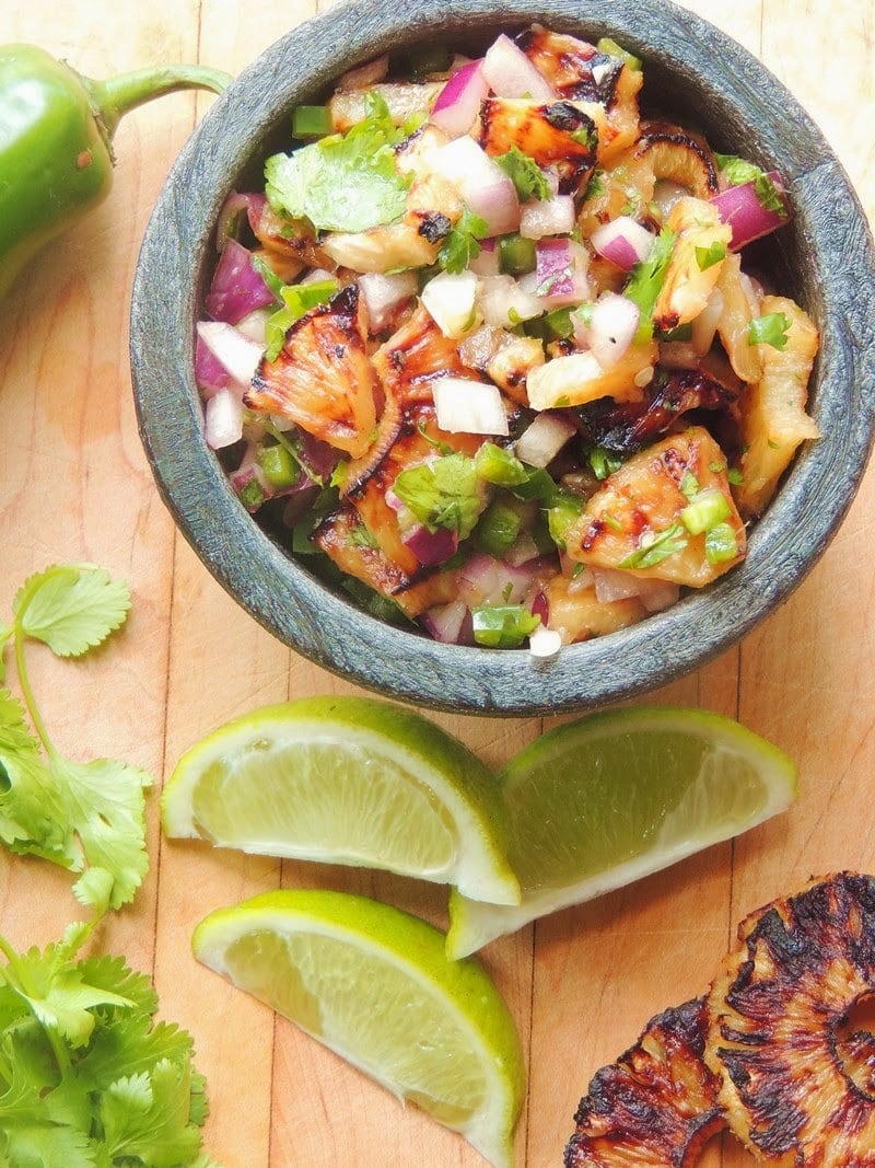 An overhead view of a small black bowl full of grilled pineapple salsa on a wooden cutting board with fresh jalapeno, cilantro, lime wedges, and a grilled pineapple ring next to the bowl.
