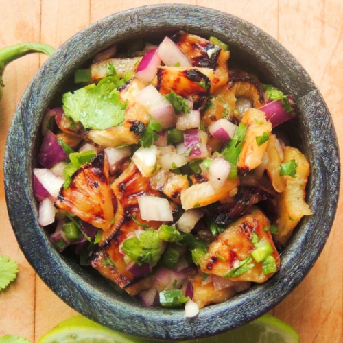 Overhead view of a black bowl full of grilled pineapple salsa on a wooden cutting board.