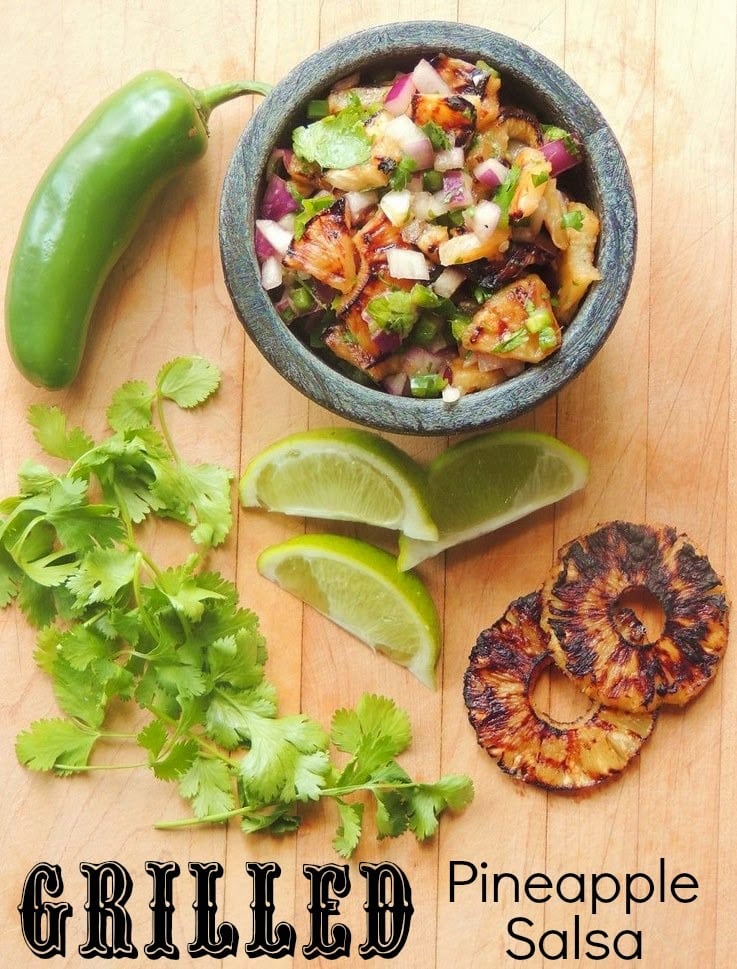 Overhead view of a black bowl full of grilled pineapple salsa on a wooden cutting board with a jalapeno, fresh cilantro, lime slices, and slices of grilled pineapple.