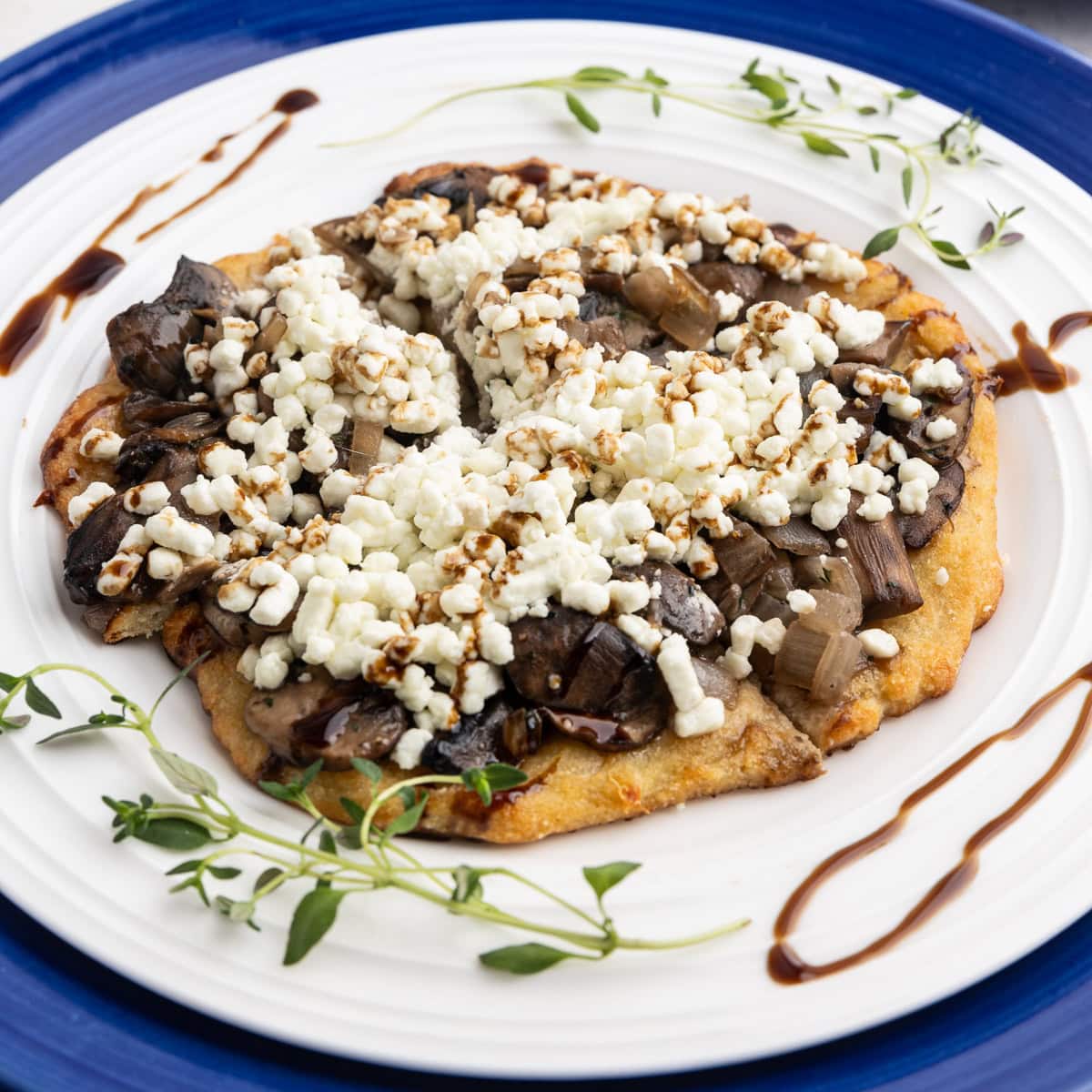 A low-carb mushroom goat cheese flat bread on a white dinner plate with sprigs of fresh time and balsamic drizzle.