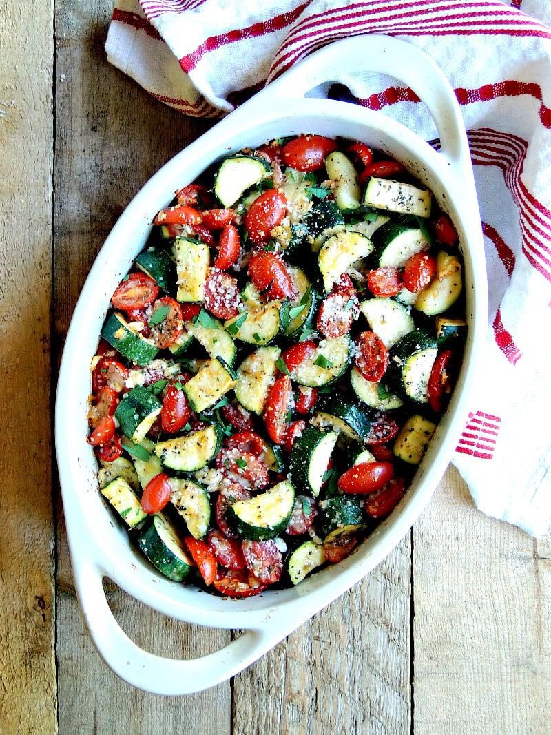 Overhead view of garlic parmesan baked zucchini and tomatoes in a white baking dish on a wooden table.