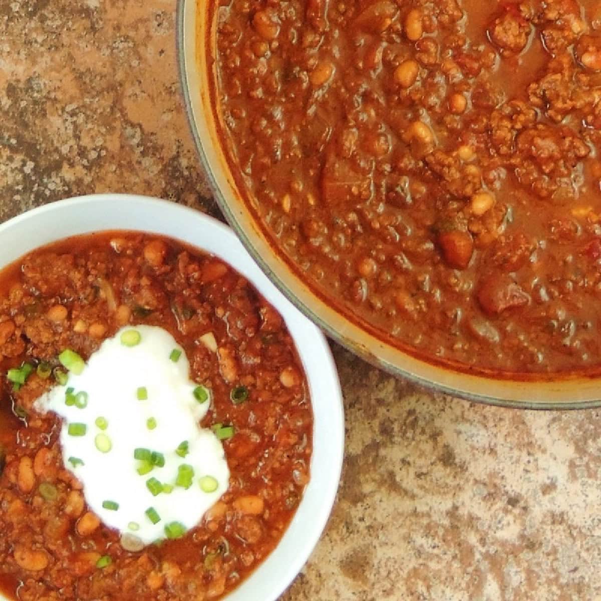 Closeup of a large pot full of pumpkin chili next to a bowl of the chili topped with sour cream.