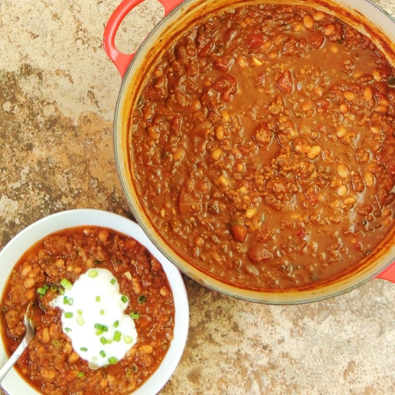 Overhead view of a large red Dutch oven full of pumpkin chili on a brown marble counter with a white bowl full of chili next to it topped with sour cream and chopped chives.